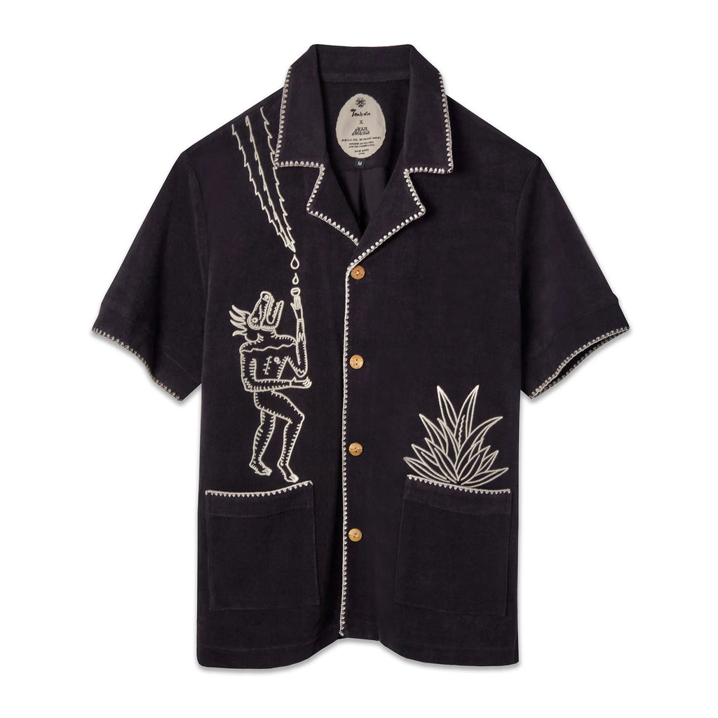A black short-sleeve terry cloth shirt with light tan embroidery. On the left chest is a stylized figure with exaggerated features, standing beneath lightning bolts and raindrops. On the right pocket is an embroidered agave plant. The shirt has wooden buttons, front patch pockets, and contrast whipstitch detailing along the collar, cuffs, and hems. ('Agua Magica' Cabana)