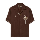 Brown short-sleeve cabana shirt with embroidered cocktail motifs, including a crescent moon and a coupe glass with garnish. The shirt has cream piping on the sleeves and “Tombolo” embroidery at the collar.