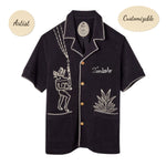 A black cabana shirt with white custom embroidery, featuring bamboo buttons and edge stitching detail, featuring 'Tombolo' across the chest. 