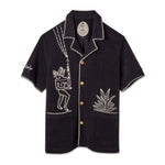 A black cabana shirt with white custom embroidery, featuring bamboo buttons and edge stitching detail, featuring 'Tombolo' on the sleeve.