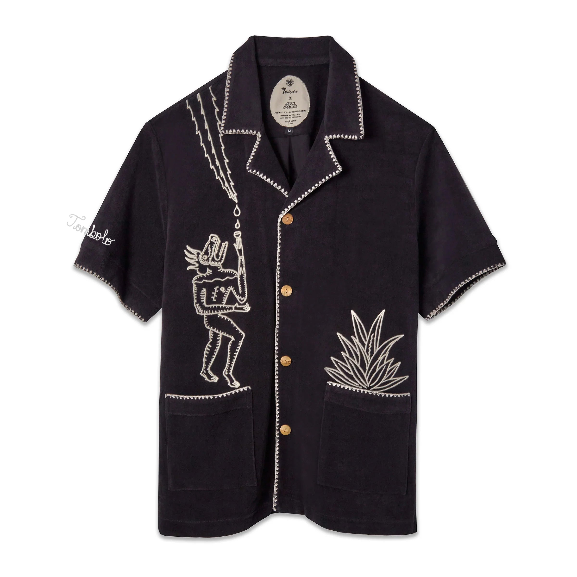 A black cabana shirt with white custom embroidery, featuring bamboo buttons and edge stitching detail, featuring 'Tombolo' on the sleeve.