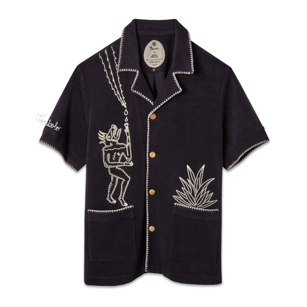 A black cabana shirt with white custom embroidery, featuring bamboo buttons and edge stitching detail, featuring 'Tombolo' on the sleeve.