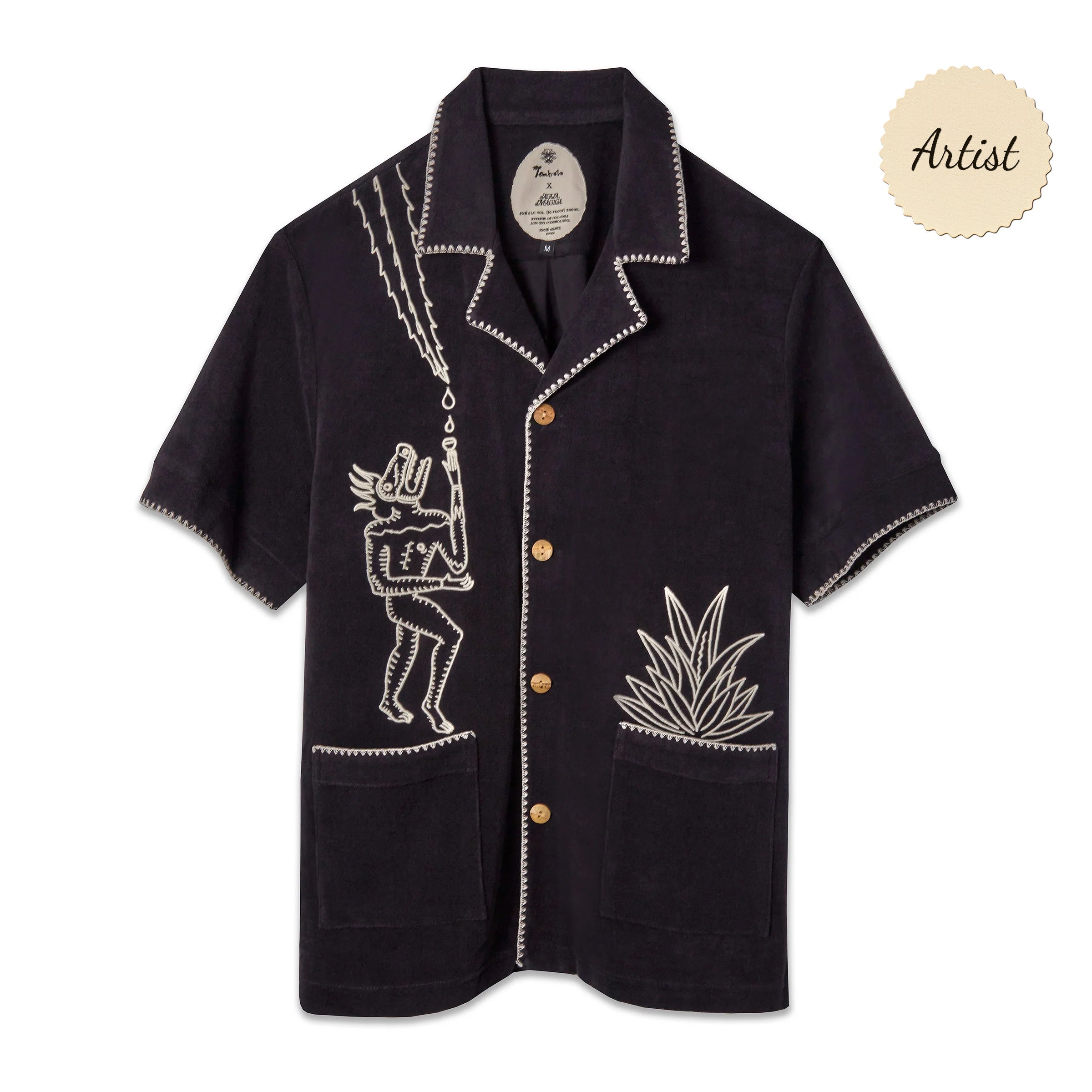 A black short-sleeve terry cloth shirt with light tan embroidery. On the left chest is a stylized figure with exaggerated features, standing beneath lightning bolts and raindrops. On the right pocket is an embroidered agave plant. The shirt has wooden buttons, front patch pockets, and contrast whipstitch detailing along the collar, cuffs, and hems. A beige sticker in the top corner reads "Artist."
