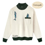 Cream knit sweater with dark green ribbed collar, cuffs, and hem. Features embroidered fish, including one hanging from a fishing line near the shoulder and two peeking out from a chest pocket. 'Tombolo' embroidered near the waist.