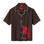 Front view of a short-sleeve, button-up shirt in dark brown with beige piping along the collar, placket, and sleeve hems. A textured red lobster applique is stitched to the lower left side, with a tan rope extending upward along the button line, mimicking the lobster's tether. The word “Tombolo” is embroidered in red inside the collar.