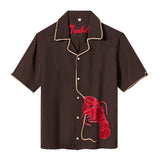 Front view of a short-sleeve, button-up shirt in dark brown with beige piping along the collar, placket, and sleeve hems. A textured red lobster applique is stitched to the lower left side, with a tan rope extending upward along the button line, mimicking the lobster's tether. The word “Tombolo” is embroidered in red inside the collar.