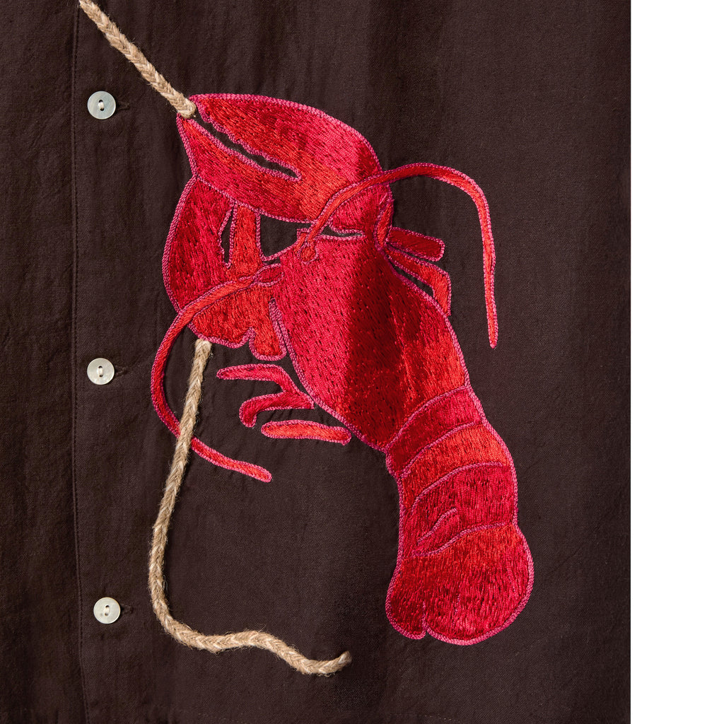 Close-up of the brown shirt featuring a detailed red lobster embroidered on the lower front. The lobster is connected to a textured, rope-like applique that runs vertically between pearlescent buttons. The embroidery has a glossy finish, creating dimension against the matte fabric.