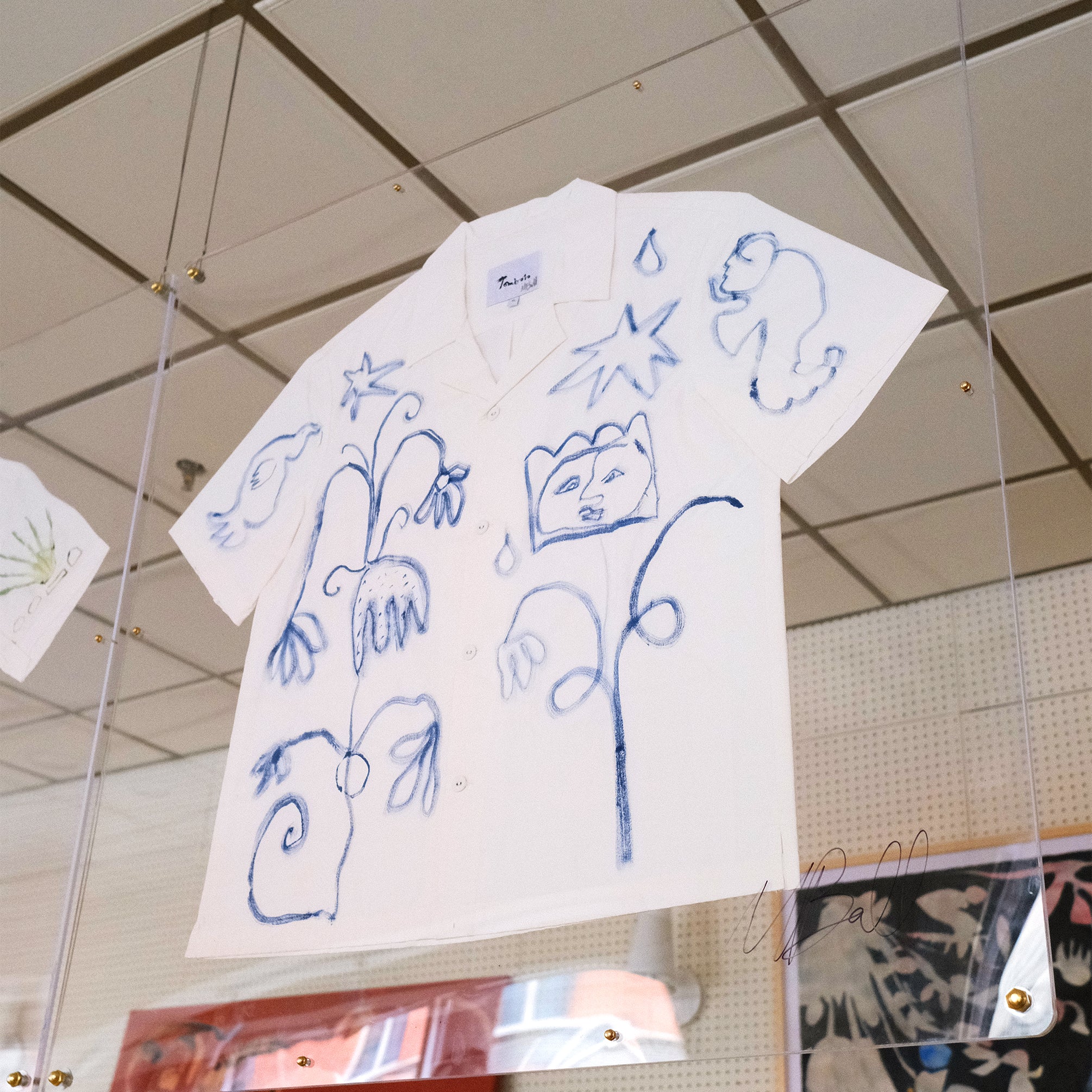 A short-sleeve white shirt with blue botanical and abstract designs, displayed inside a glass case.