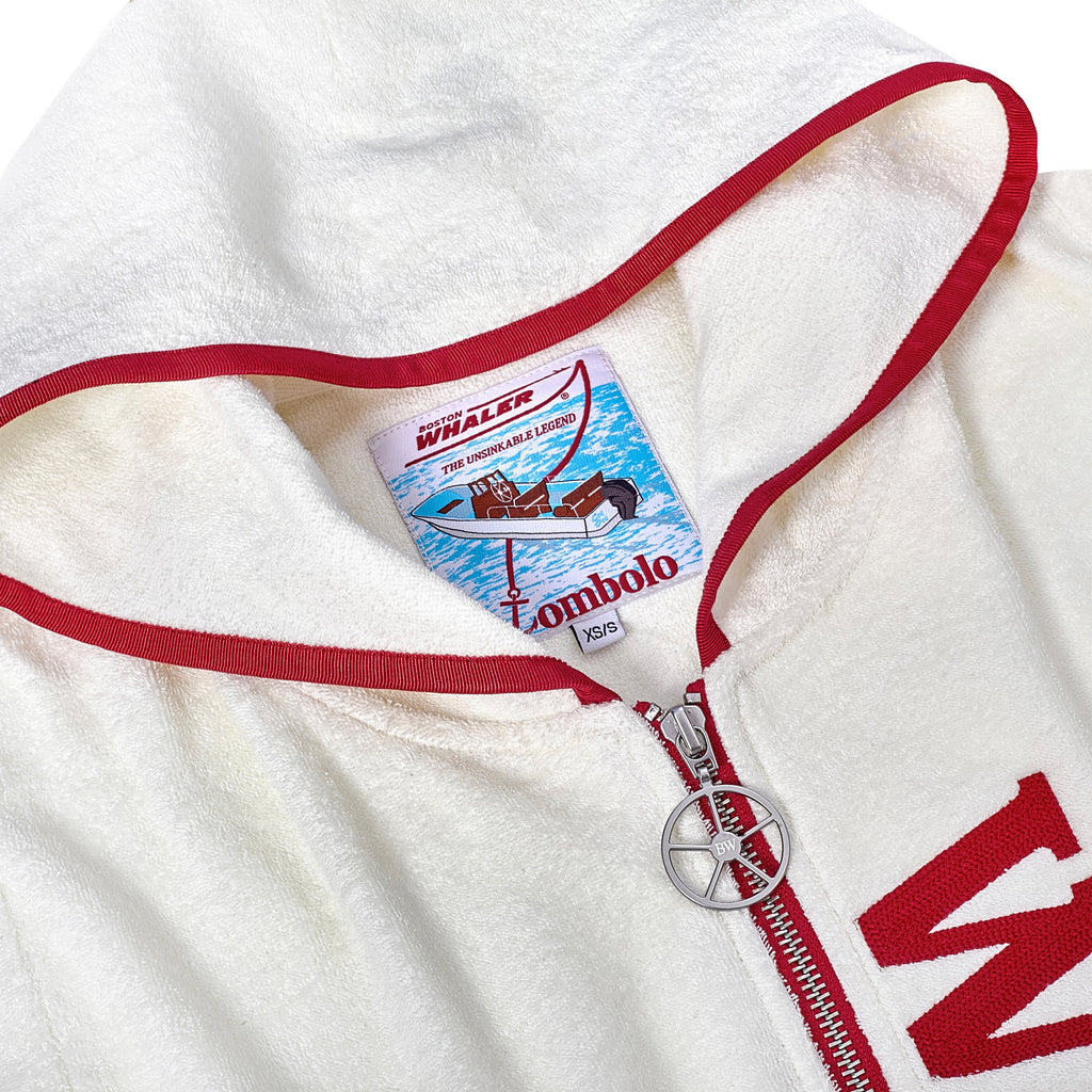 Close-up of a white terry cloth hooded beach towel top with red trim, showing the interior label with a Boston Whaler boat illustration and 'Tombolo' branding. A silver ship wheel zipper pull is visible below the label.