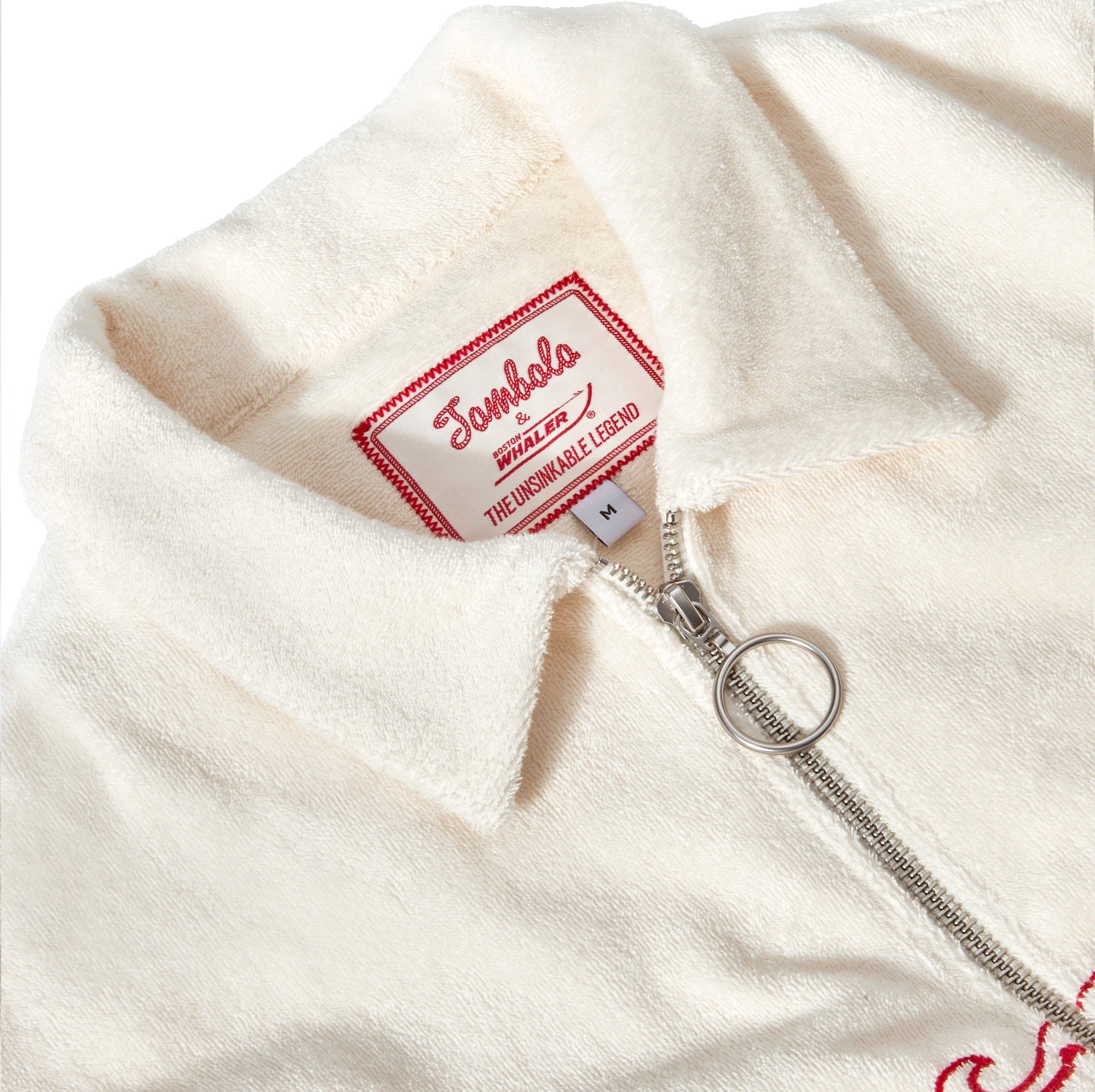 Close-up of the collar and zipper of an off-white terry cloth shirt. A silver zipper with a circular pull is partially unzipped, revealing a woven label that reads: “Tombolo x Boston Whaler – The Unsinkable Legend.” The label is red and white with a stitched border, and the fabric has a soft, textured appearance.