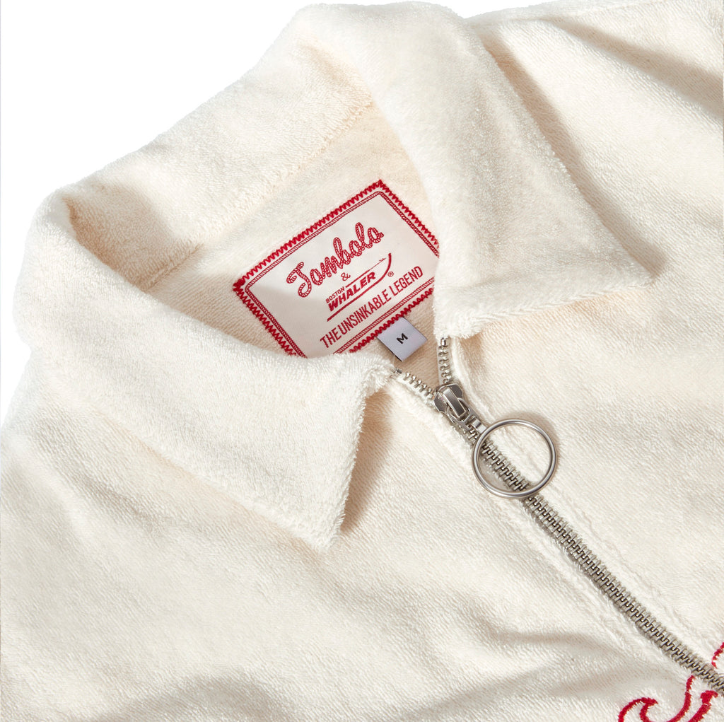 Close-up of the collar and zipper of an off-white terry cloth shirt. A silver zipper with a circular pull is partially unzipped, revealing a woven label that reads: “Tombolo x Boston Whaler – The Unsinkable Legend.” The label is red and white with a stitched border, and the fabric has a soft, textured appearance.
