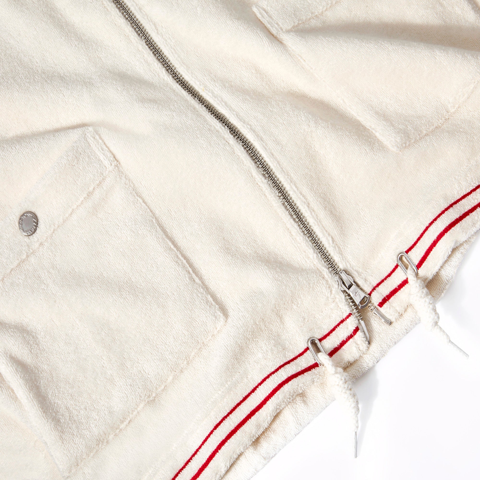 Close-up of the lower front portion of the white terry cloth shirt. Visible details include two patch pockets with snap buttons, a silver zipper down the center, and a drawstring hem with metal grommets. The hem is accented with two parallel red stripes.