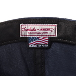 Close-up product image of the 'Boston-Whaler' 5-Panel Souvenir Cap's interior tag with Tombolo & Boston Whaler logos and 'the unsinkable legend' text below that with a made in the USA tag as well.
