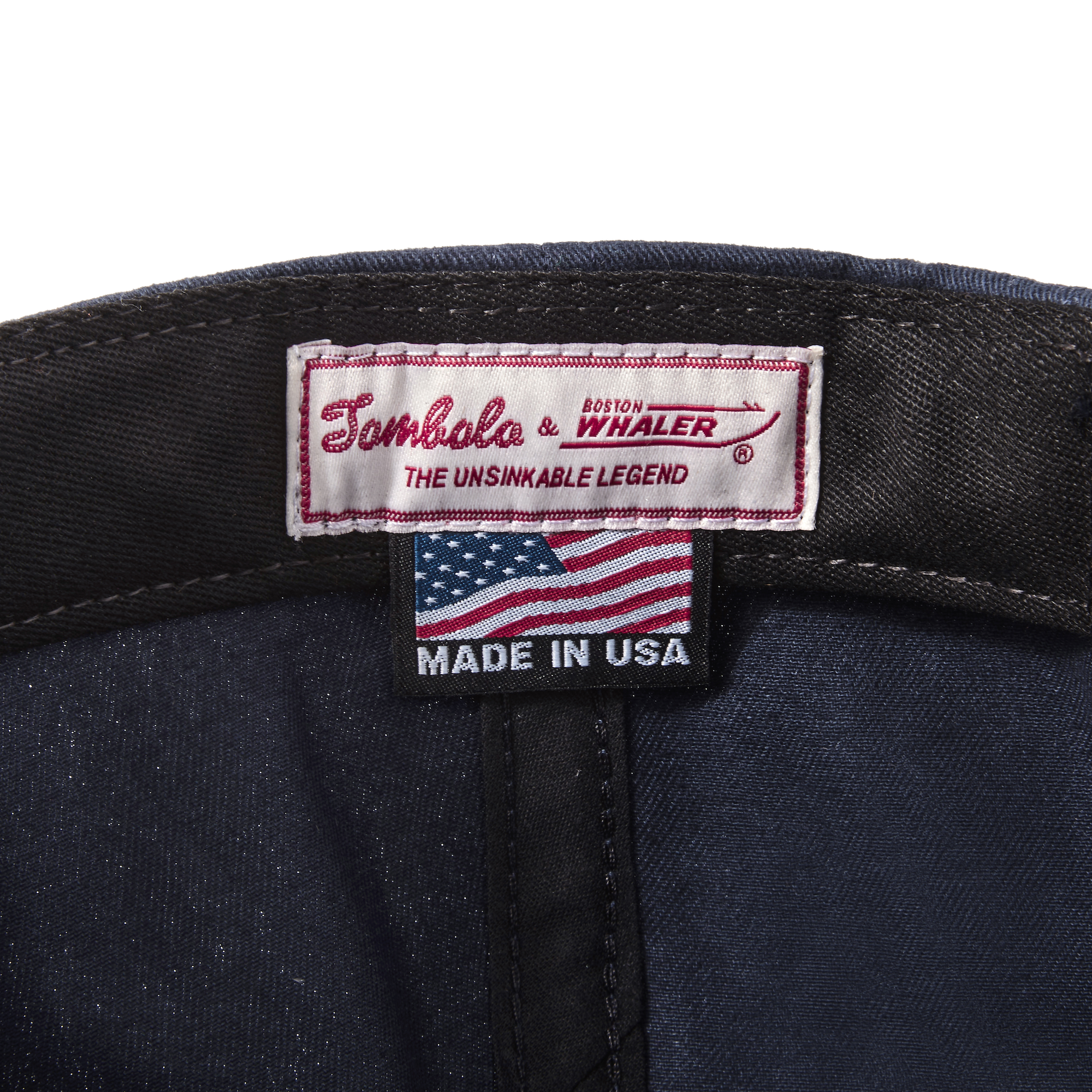 Close-up product image of the 'Boston-Whaler' 5-Panel Souvenir Cap's interior tag with Tombolo & Boston Whaler logos and 'the unsinkable legend' text below that with a made in the USA tag as well.