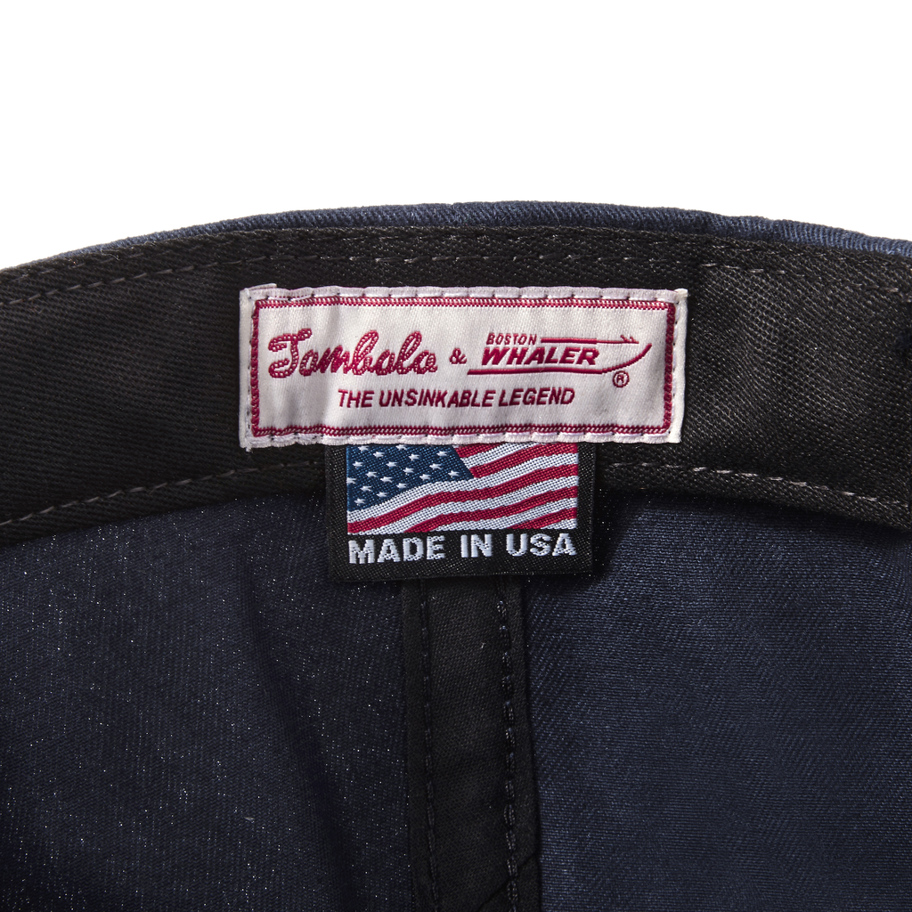 Close-up product image of the 'Boston-Whaler' 5-Panel Souvenir Cap's interior tag with Tombolo & Boston Whaler logos and 'the unsinkable legend' text below that with a made in the USA tag as well.