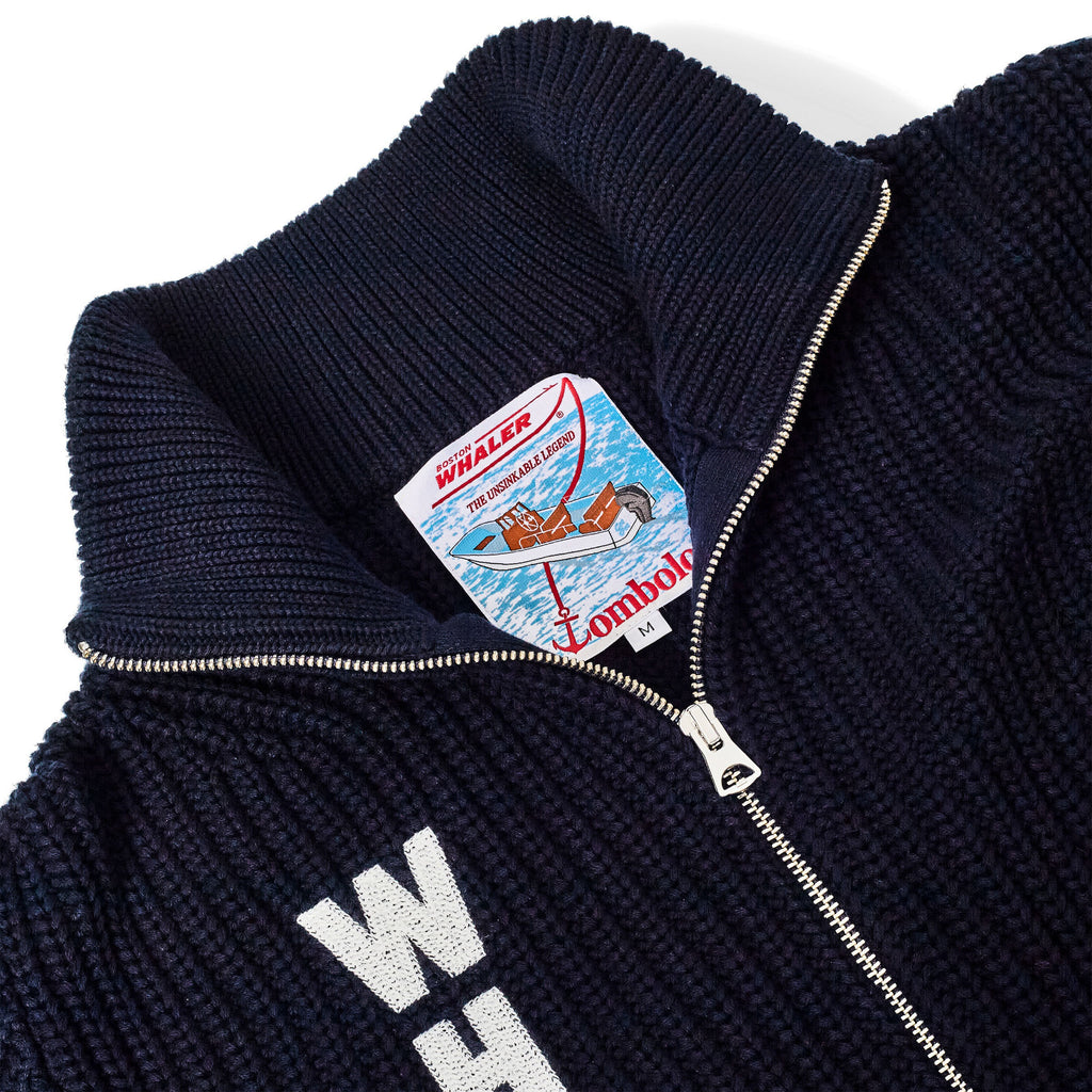 Close-up of a navy knit zip-up cardigan with textured ribbing and vertical 'WHALER' lettering in white. The inside label features an illustrated Boston Whaler boat above Tombolo branding.