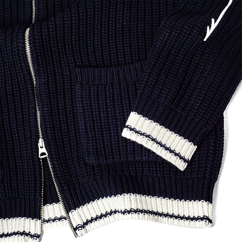 Detail of a navy ribbed knit cardigan showing front pockets, a silver zipper, and striped white-and-navy trim at the hem and cuffs.
