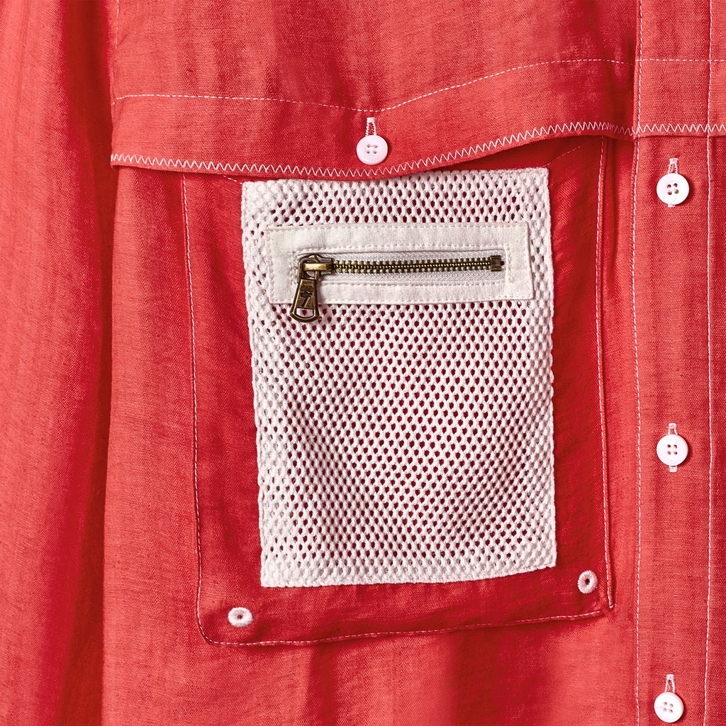 Close-up of a red shirt featuring a white mesh pocket with a small brass zipper. The pocket is stitched onto the chest and surrounded by white contrast stitching and white buttons.