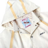 Close-up of a cream hooded jacket with tan trim and silver snap buttons. The inside features mesh lining and a colorful interior tag with an illustrated Boston Whaler boat, reading 'The Unsinkable Legend' and 'Tombolo.'