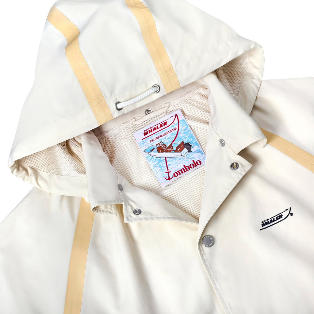 Close-up of a cream hooded jacket with tan trim and silver snap buttons. The inside features mesh lining and a colorful interior tag with an illustrated Boston Whaler boat, reading 'The Unsinkable Legend' and 'Tombolo.'