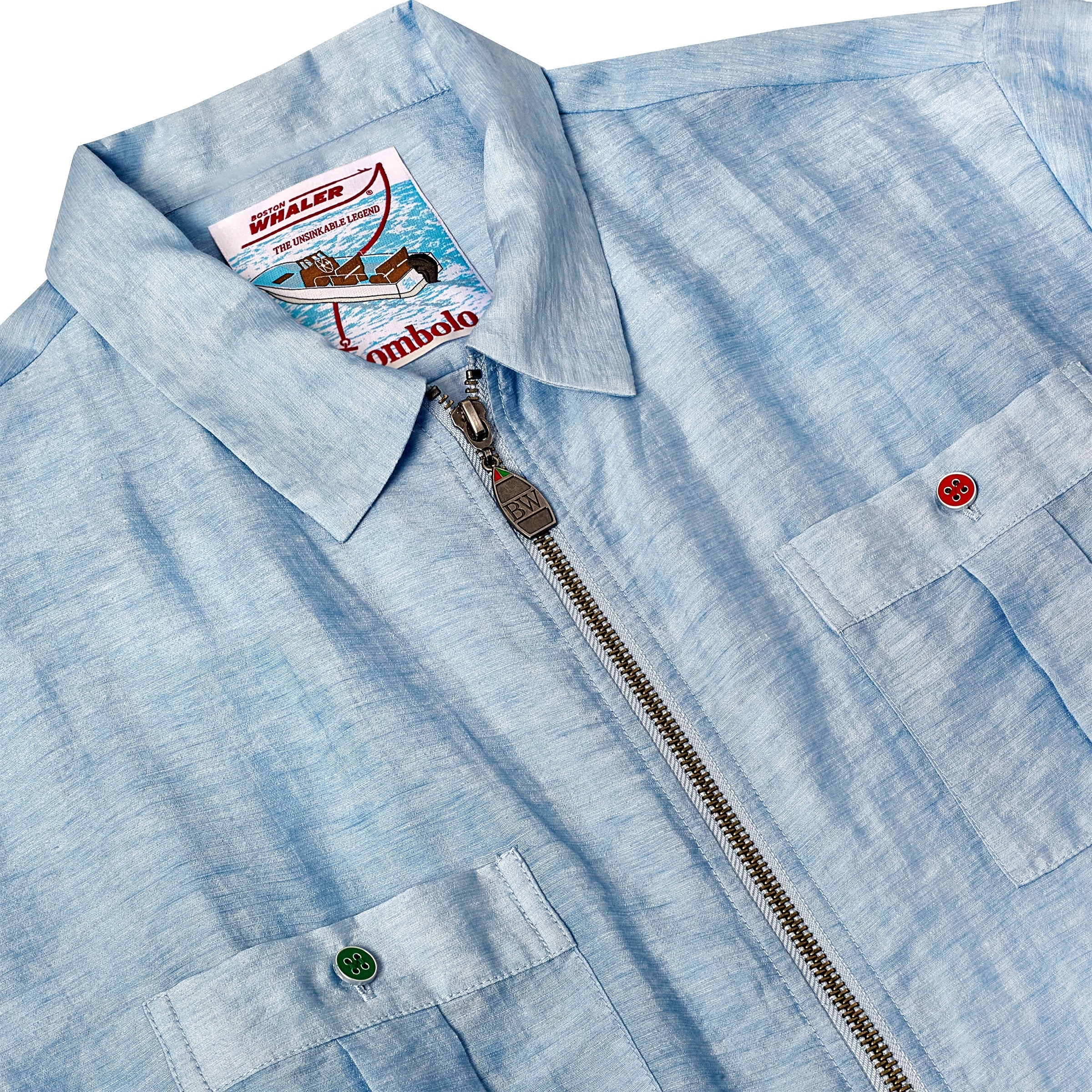 Close-up of a light blue short-sleeve shirt with a silver zipper and two front pockets. One pocket has a green button and the other has a red button. The label inside the collar features a Boston Whaler boat illustration and branding from Tombolo.