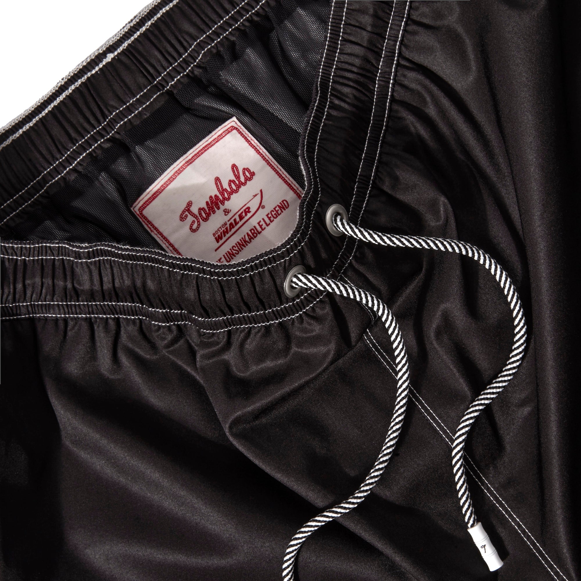 Close up product image of the 'boston whaler' swim trunks' interior label and metal eyelet stoppers on white and black twist drawstrings.