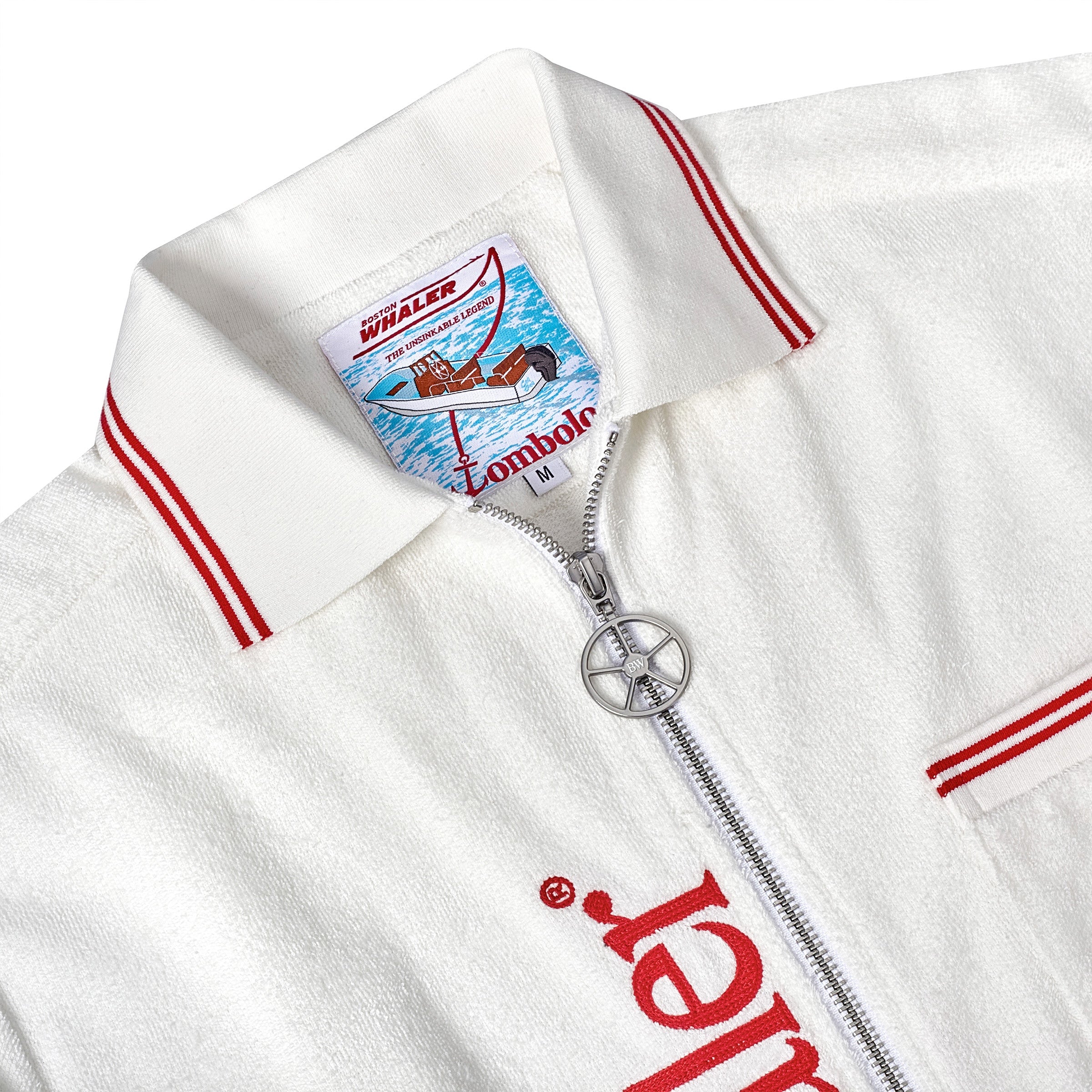 Close-up of a white terry cloth cabana with red trim, showing the interior label with a Boston Whaler boat illustration and 'Tombolo' branding. A silver ship wheel zipper pull is visible below the label.