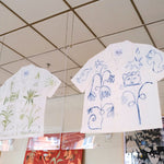 A short-sleeve white shirt with green botanical and abstract designs, and a white shirt with blue abstract designs, displayed inside two glass cases. 