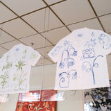 A short-sleeve white shirt with green botanical and abstract designs, and a white shirt with blue abstract designs, displayed inside two glass cases. 