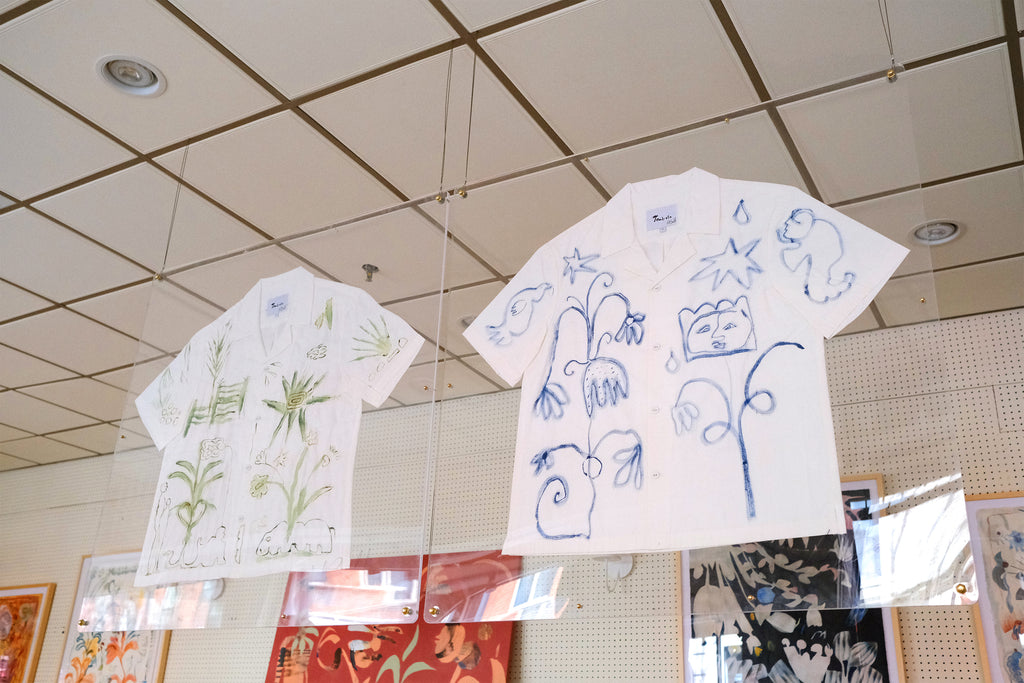 A short-sleeve white shirt with green botanical and abstract designs, and a white shirt with blue abstract designs, displayed inside two glass cases. 