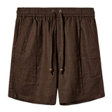 A pair of dark brown linen shorts with a relaxed fit. They feature an elastic waistband with a matching drawstring that includes wooden bead tips. The shorts have two side pockets and subtle stitching details throughout, offering a breezy, casual look.