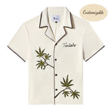 Short-sleeve cream cabana shirt with brown trim, embroidered green leaves, and the word 'Tombolo' stitched on the chest. Shirt is labeled customizable.