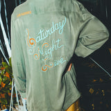 Woman wearing a green shirt with 'Saturday Night Live' text on the back, standing in front of a bar cabinet. 
