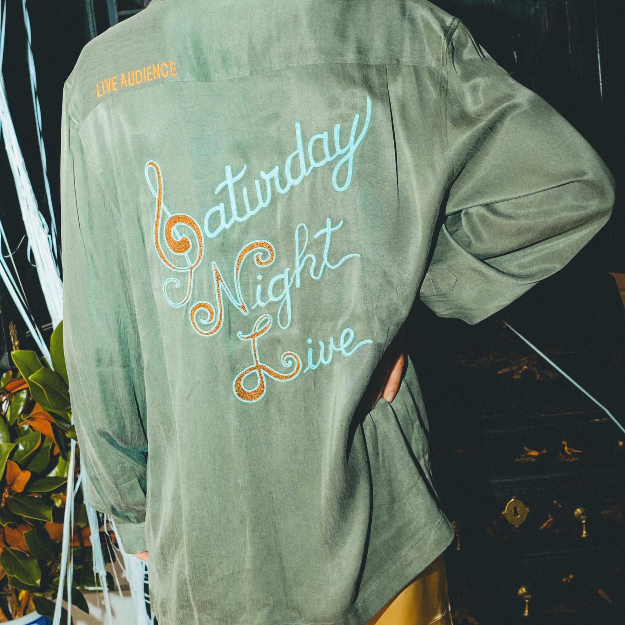 Woman wearing a green shirt with 'Saturday Night Live' text on the back, standing in front of a bar cabinet. 