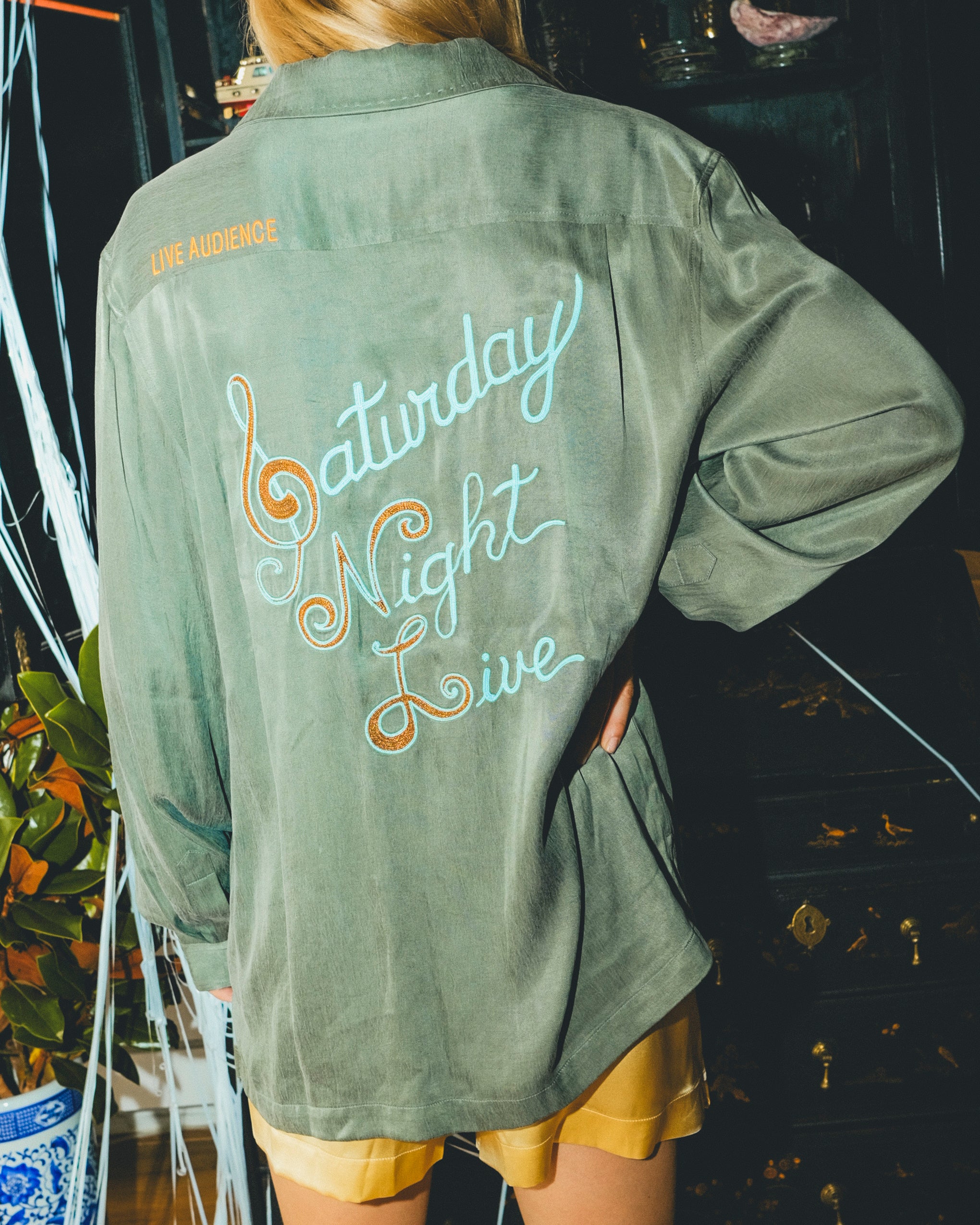 Woman wearing a green shirt with 'Saturday Night Live' text on the back, standing in front of a bar cabinet. 
