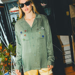 Woman wearing a green SNL shirt with colorful buttons in front of a bar cabinet. 