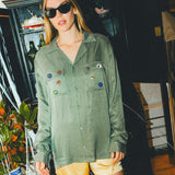 Woman wearing a green SNL shirt with colorful buttons in front of a bar cabinet. 
