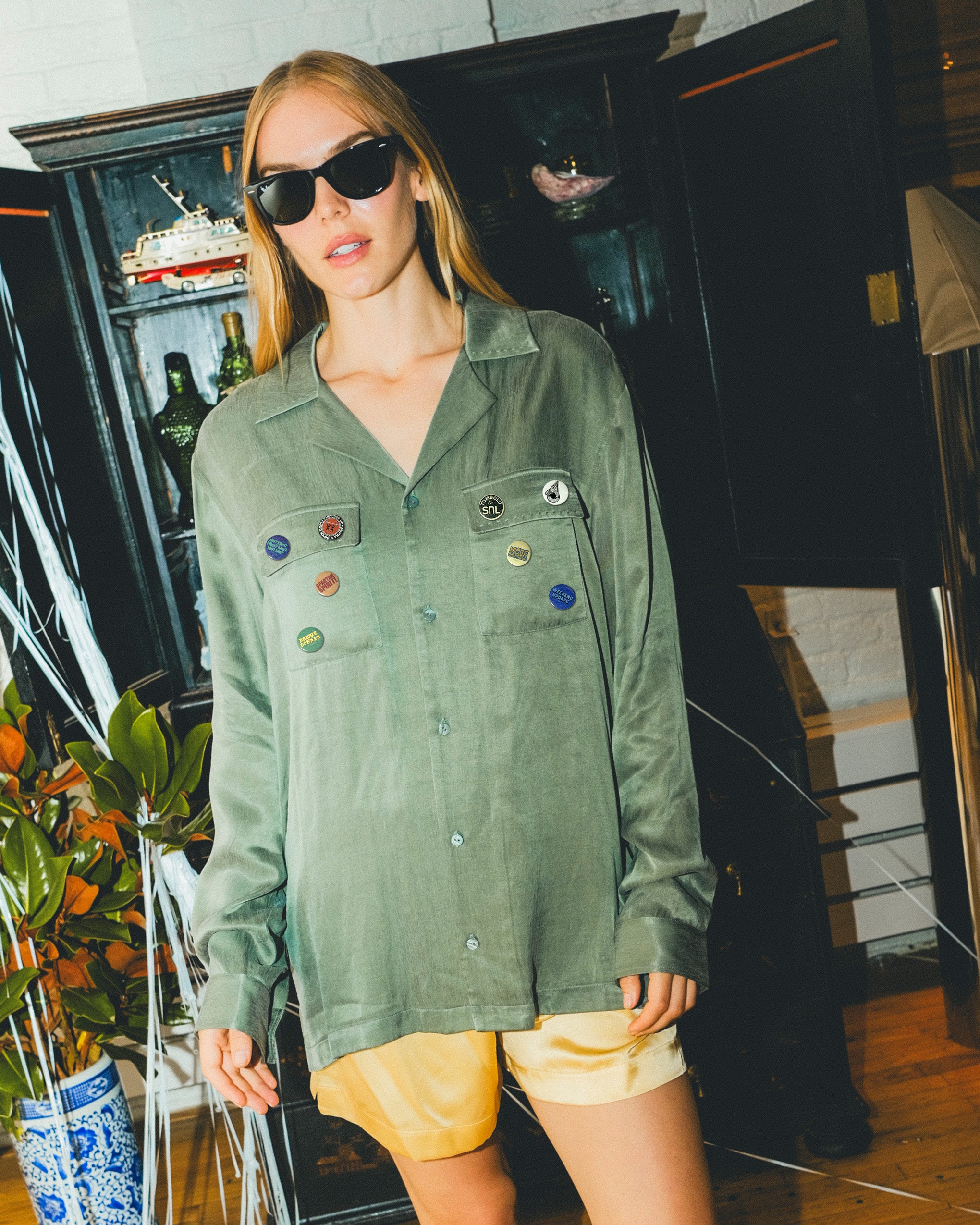 Woman wearing a green SNL shirt with colorful buttons in front of a bar cabinet. 
