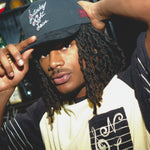 Man wearing a black cap with Saturday Night Live embroidery and a white shirt with musical notes.