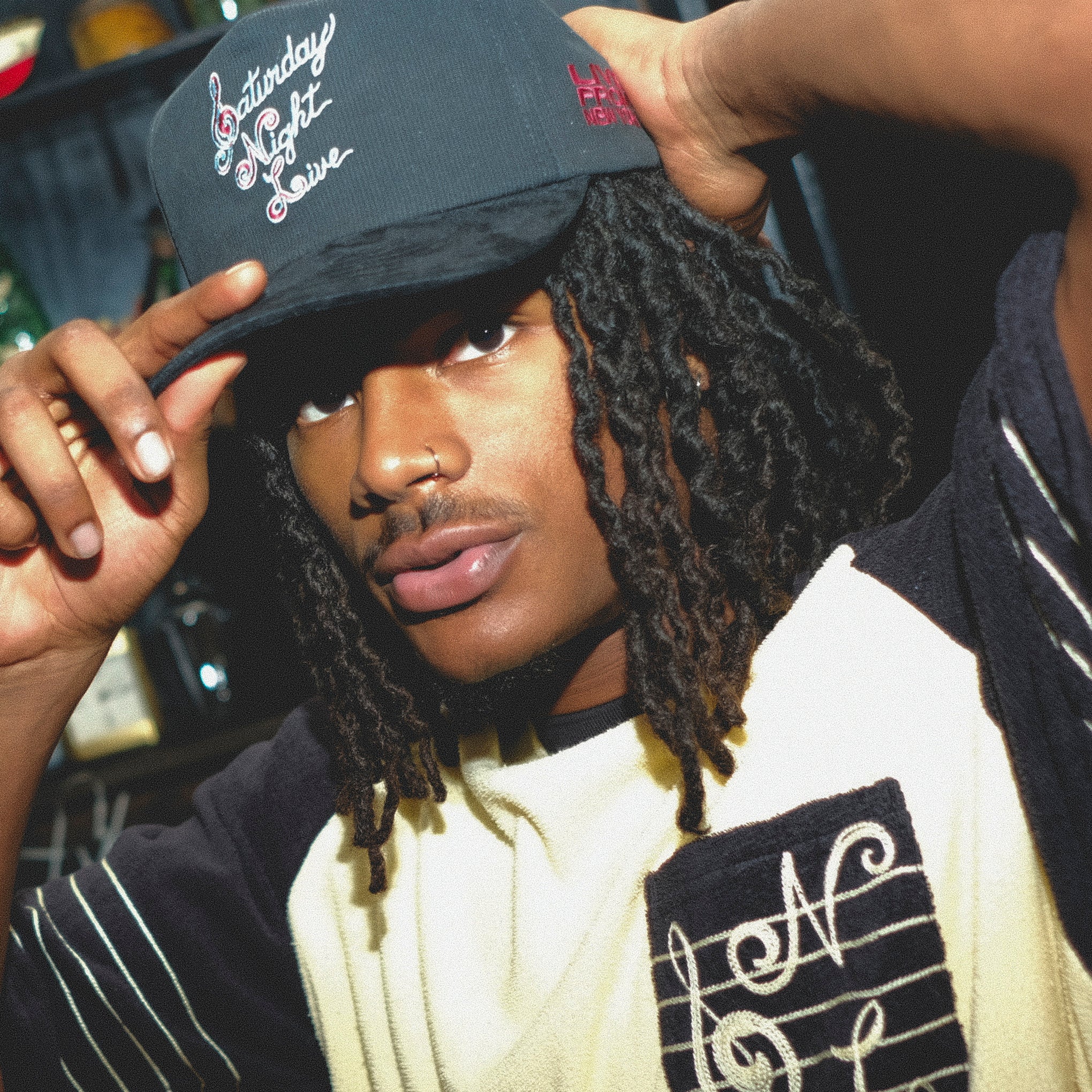 Man wearing a black cap with Saturday Night Live embroidery and a white shirt with musical notes.