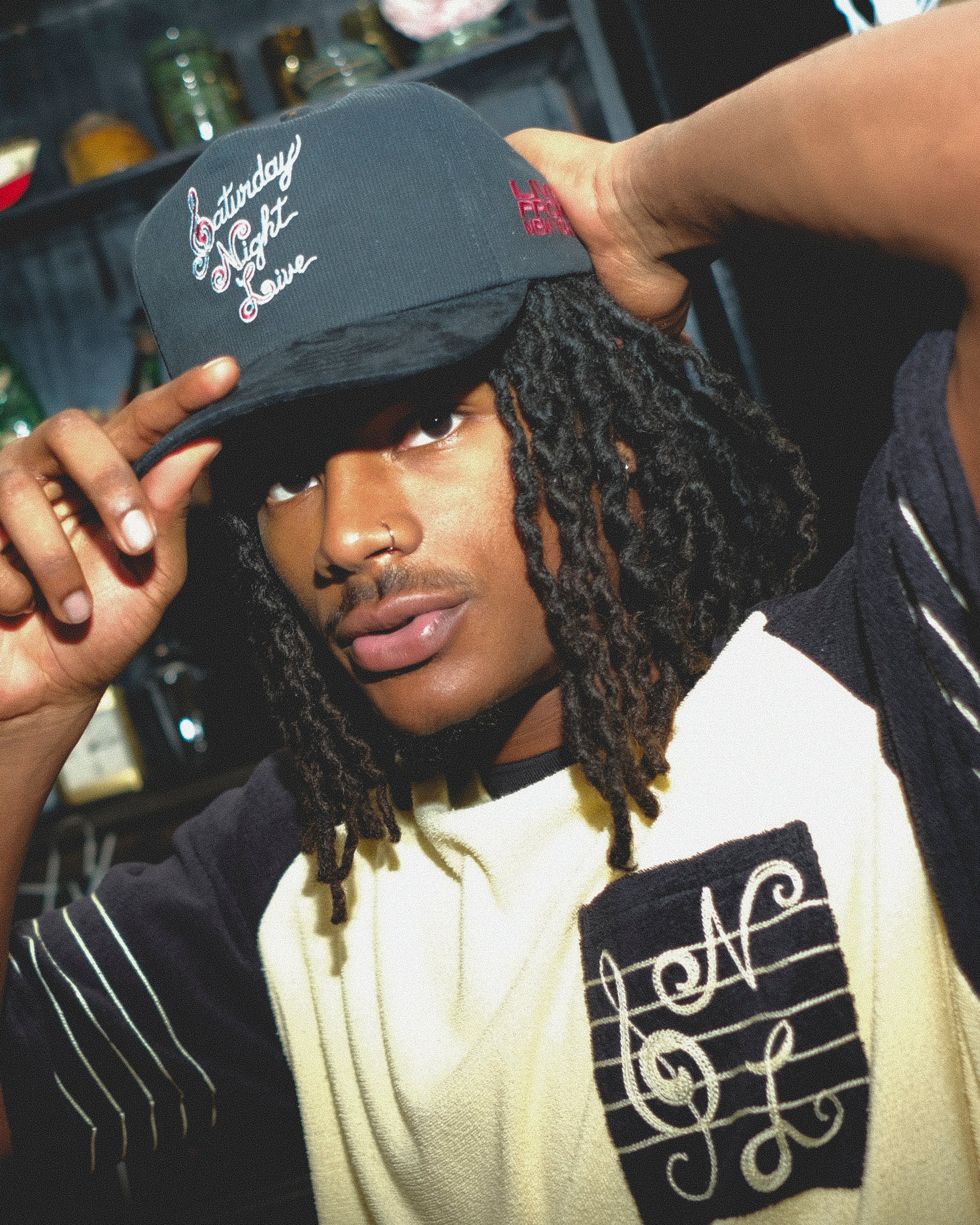 Man wearing a black cap with Saturday Night Live embroidery and a white shirt with musical notes.