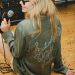 Person wearing a green shirt with 'Saturday Night Live' text on the back, sitting on the floor with a microphone.