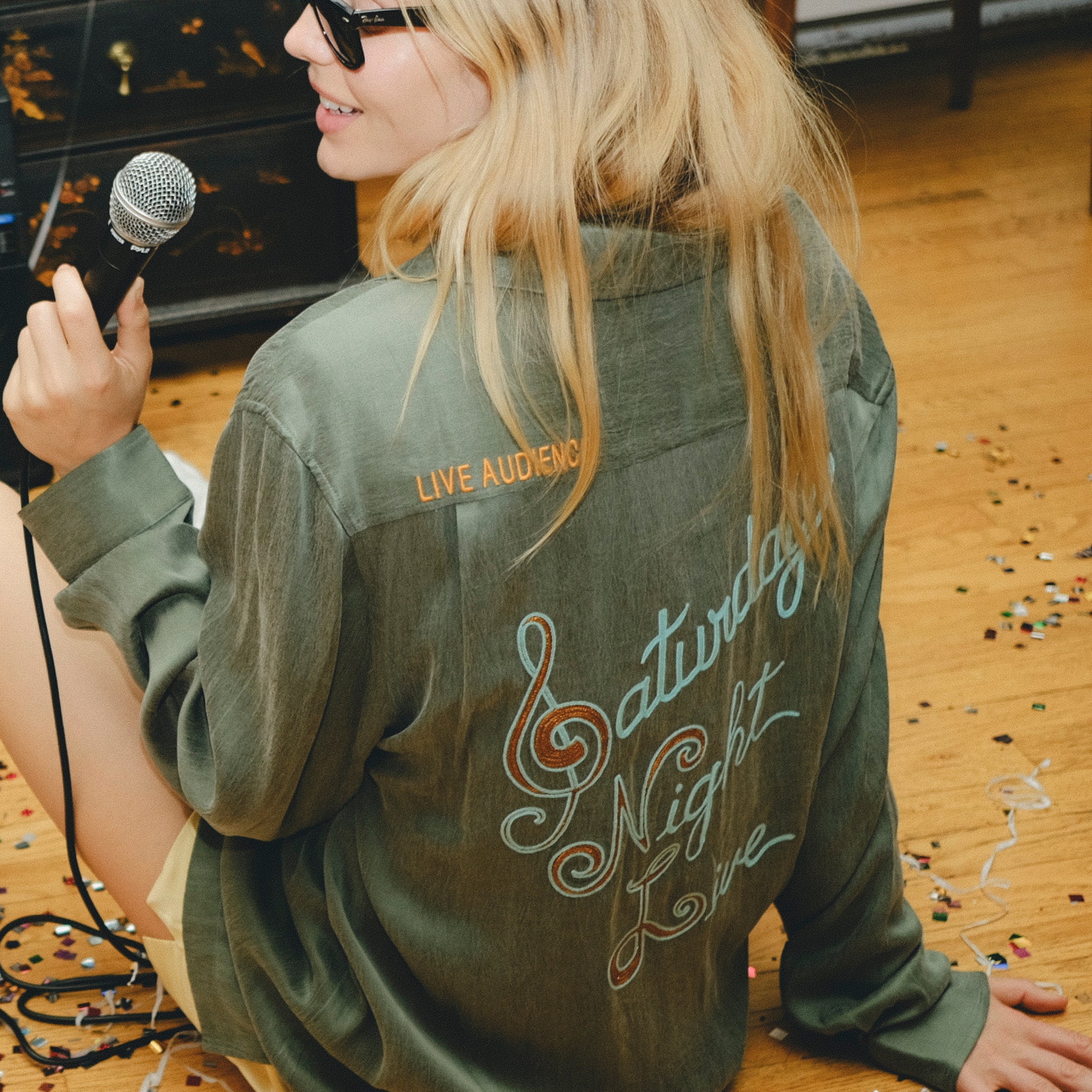 Person wearing a green shirt with 'Saturday Night Live' text on the back, sitting on the floor with a microphone.
