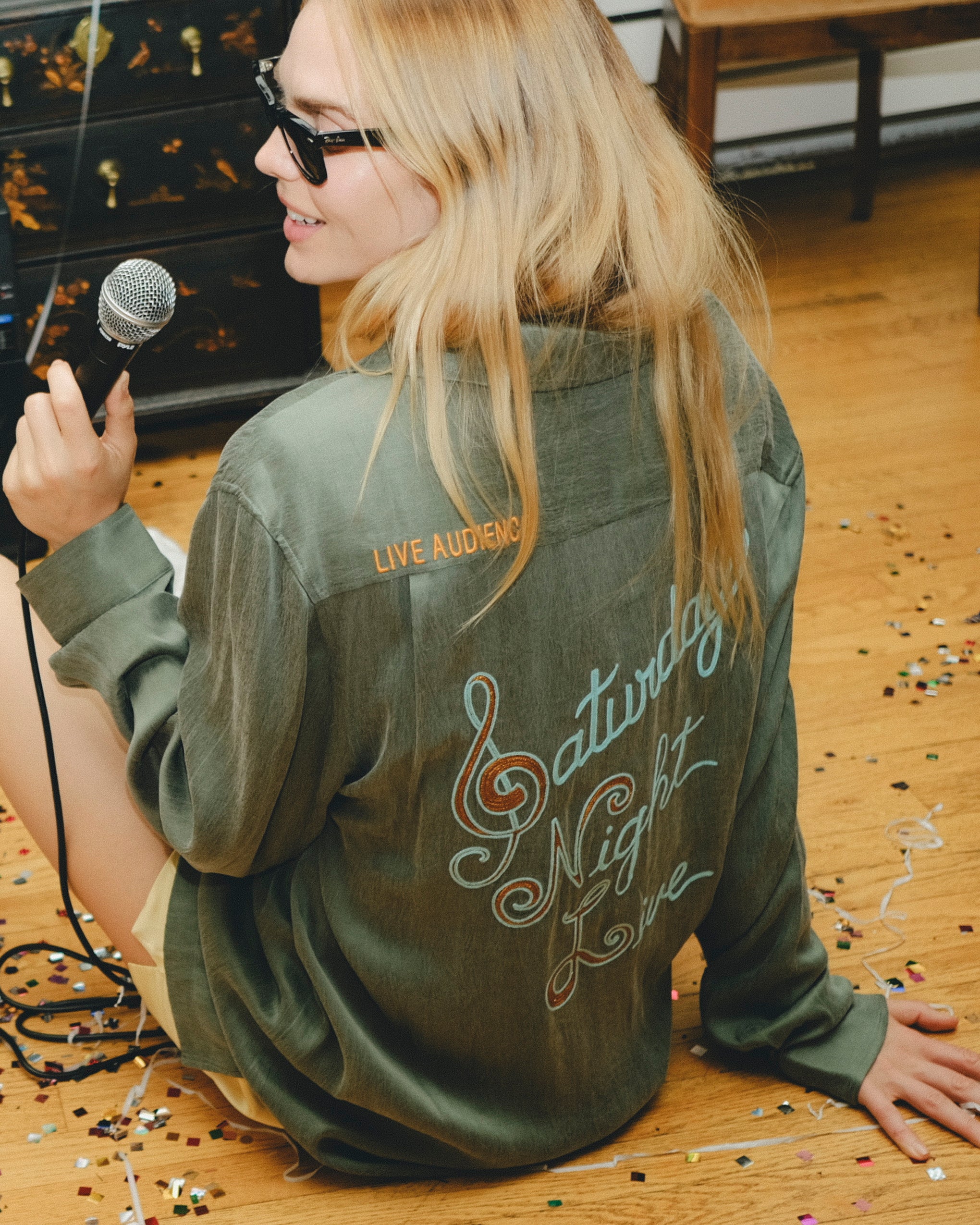 Person wearing a green shirt with 'Saturday Night Live' text on the back, sitting on the floor with a microphone.