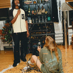 Two people with microphones in a room with a large cabinet filled with bottles and decorative items, wearing SNL products. 