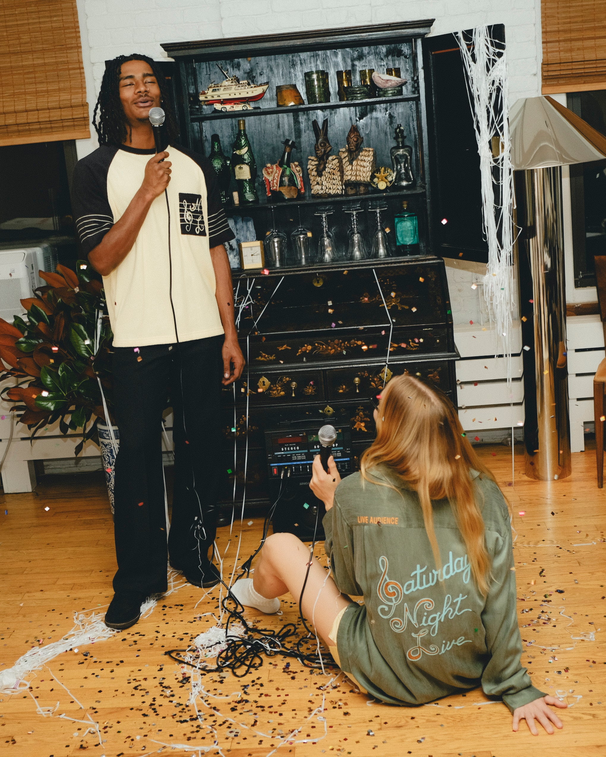 Two people with microphones in a room with a large cabinet filled with bottles and decorative items, wearing SNL products. 