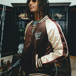 Man wearing SNL brown satin souvenir jacket with cream sleeves and striped trim.