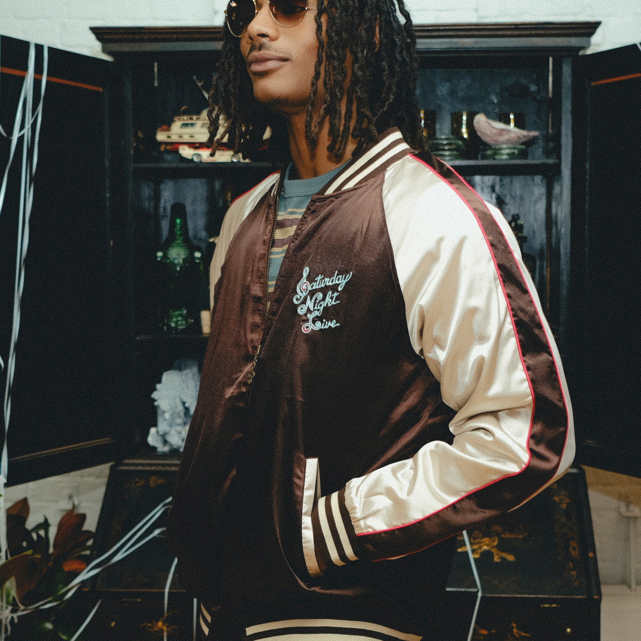 Man wearing SNL brown satin souvenir jacket with cream sleeves and striped trim.