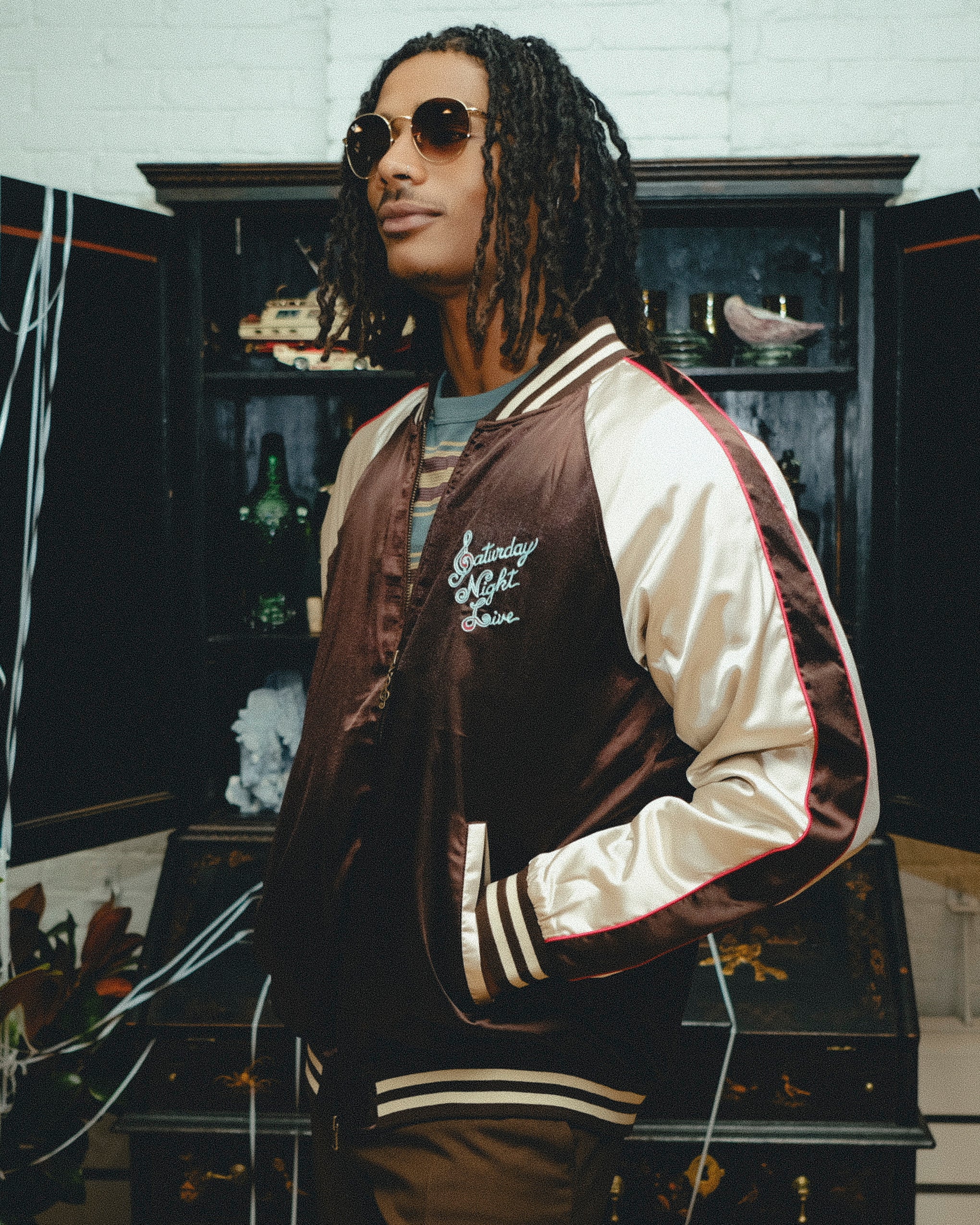 Man wearing SNL brown satin souvenir jacket with cream sleeves and striped trim.