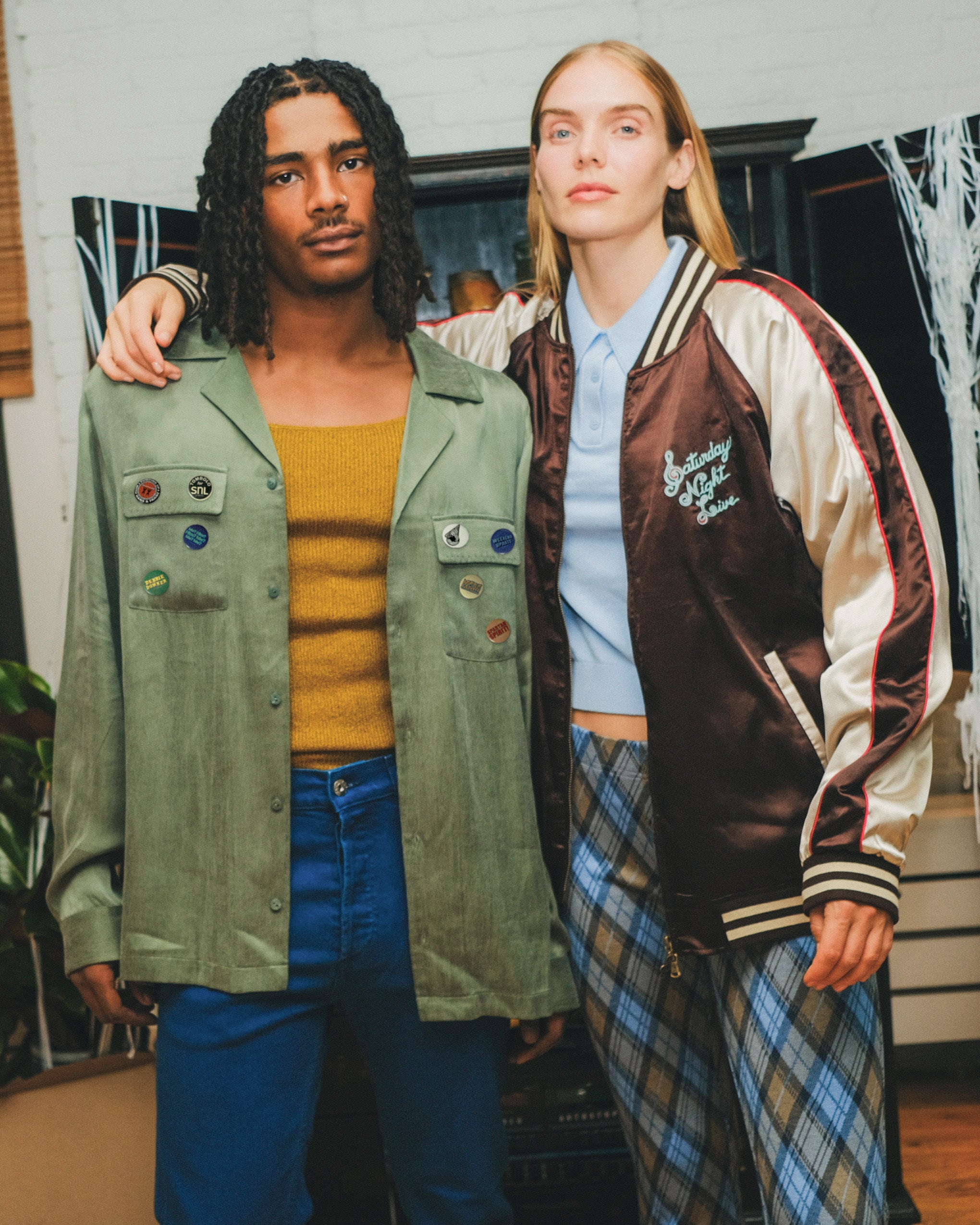 Two models standing together, wearing the SNL Long Sleeve 1976 shirt and the Souvenir Jacket. 